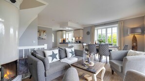 A cozy living room with a fireplace, grey sofas with star pillows, a wooden coffee table, and a dining area in the background. - Lifestyle Unter Reet, Strand & Meer um die Ecke (Wenningstedt-Braderup (Sylt))