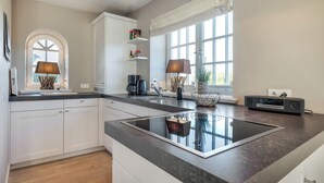 A modern kitchen with white cabinets, a black countertop, and a built-in induction cooktop. There are two lamps on the countertop, a coffee maker, and a radio. - Lifestyle Unter Reet, Strand & Meer um die Ecke. Ihr Optimales Urlaubs-domizil (Wenningstedt-Braderup (Sylt))