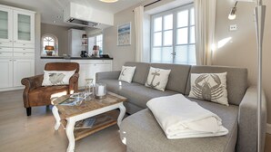 A living room with a sofa, armchair, coffee table, and a window with curtains. - Lifestyle Unter Reet, Strand & Meer um die Ecke. Ihr Optimales Urlaubs-domizil (Wenningstedt-Braderup (Sylt))