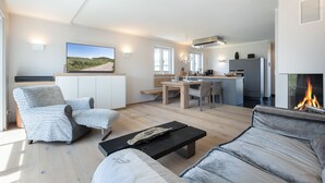 Ein modernes Wohnzimmer mit einer Couch, einem Couchtisch und einem Fernseher. Ein Kamin mit offenem Feuer ist zu sehen.