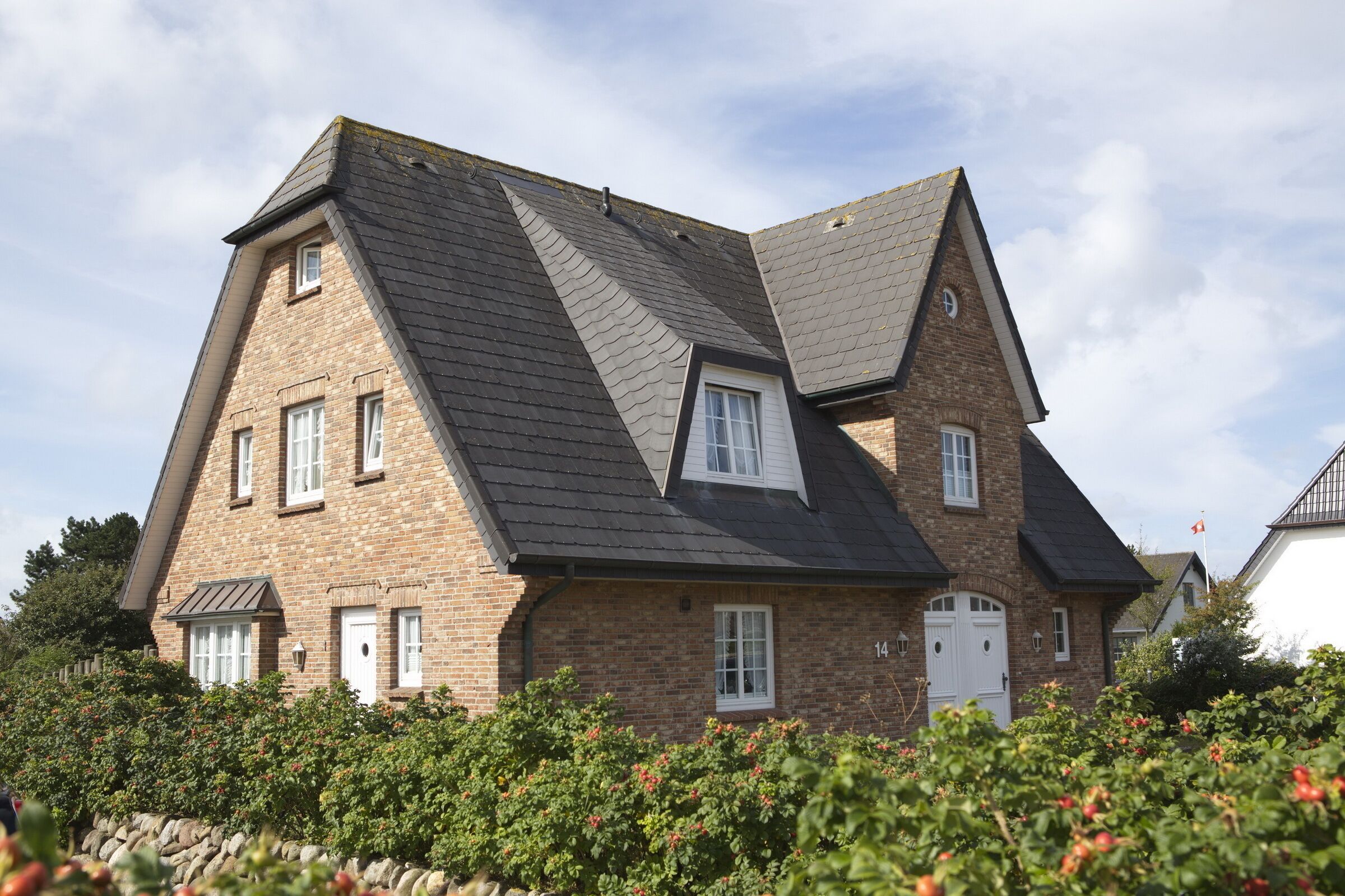 A two-story house with a steeply pitched roof, brick exterior, and a stone wall in the foreground.