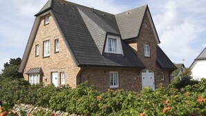 A two-story house with a steeply pitched roof, brick exterior, and a stone wall in the foreground.
