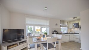 A modern kitchen with white cabinetry, a dining table with four chairs, a flat-screen TV, and a window with blinds.
