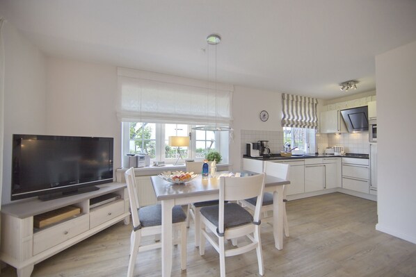 A modern kitchen with white cabinetry, a dining table with four chairs, a flat-screen TV, and a window with blinds. - Idylle & Komfort im Einklang (Wenningstedt-Braderup (Sylt))