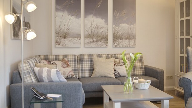 A modern living room with a grey sofa, a glass coffee table, and a floor lamp. There are three framed pictures on the wall and a vase with flowers on the table.