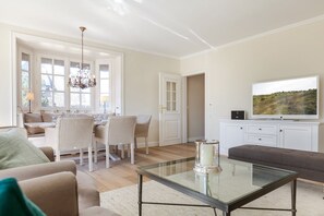 A spacious living room with a glass coffee table, a television, and a dining area with chairs and a chandelier. - Das Somerhüs Wird Ihnen Nicht nur bei Warmen Temperaturen Urlaubsfreude Bereiten (Wenningstedt-Braderup (Sylt))