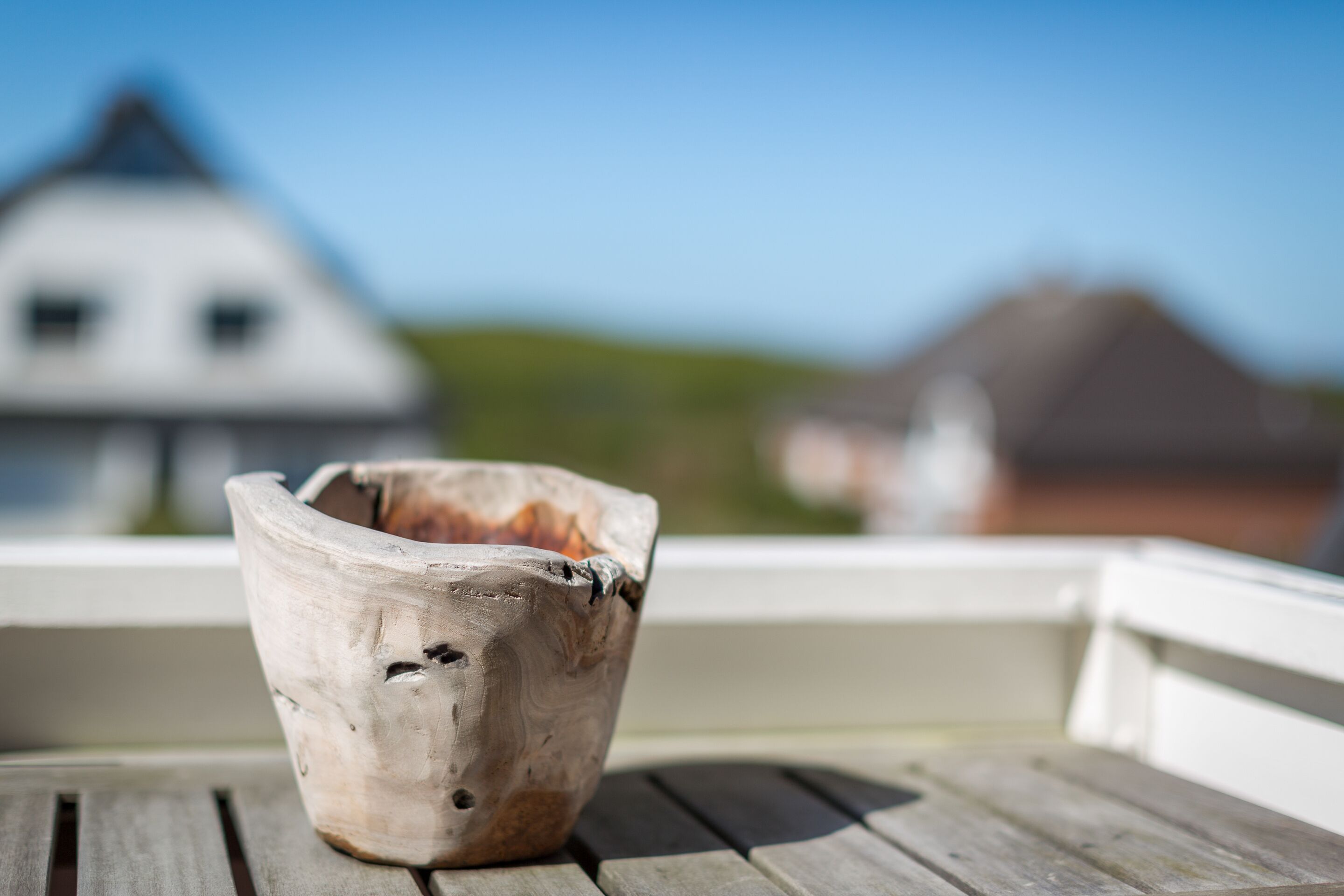 A rustic wooden table with a weathered pot on it, set against a backdrop of a white building with a triangular roof and a clear blue sky.