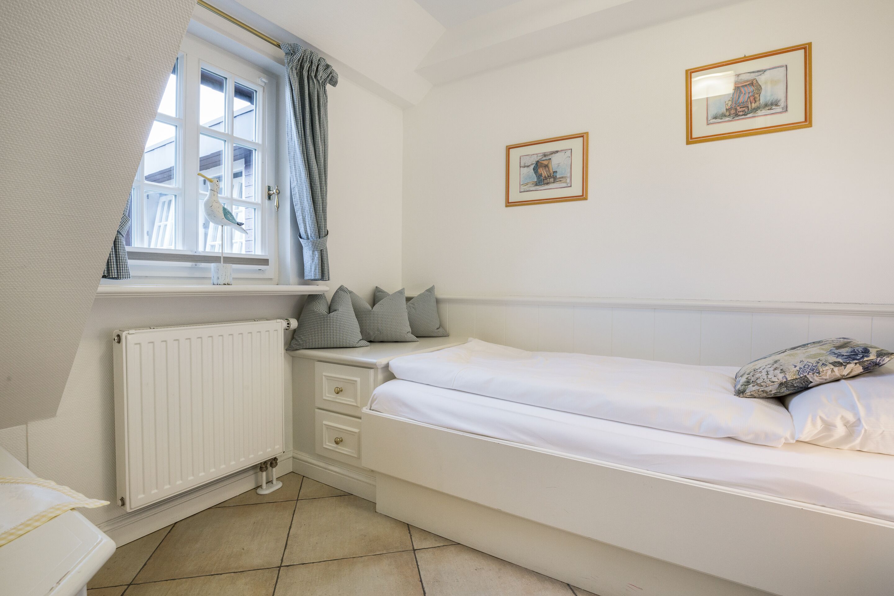 Een slaapkamer met een enkel bed, een raam met gordijnen, een radiator en twee prenten in een lijst aan de muur.