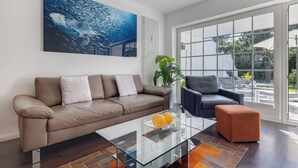 A modern living room with a brown leather sofa, a glass coffee table with oranges, a grey armchair, and a large window with a view of the outdoors.