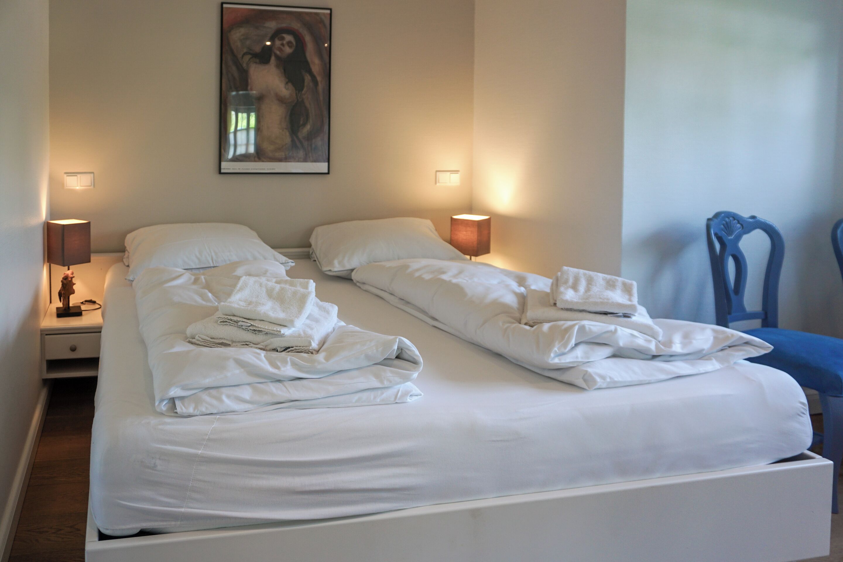 Un lit double avec du linge de lit et des serviettes blanches, deux lampes de chevet et un tableau au mur.