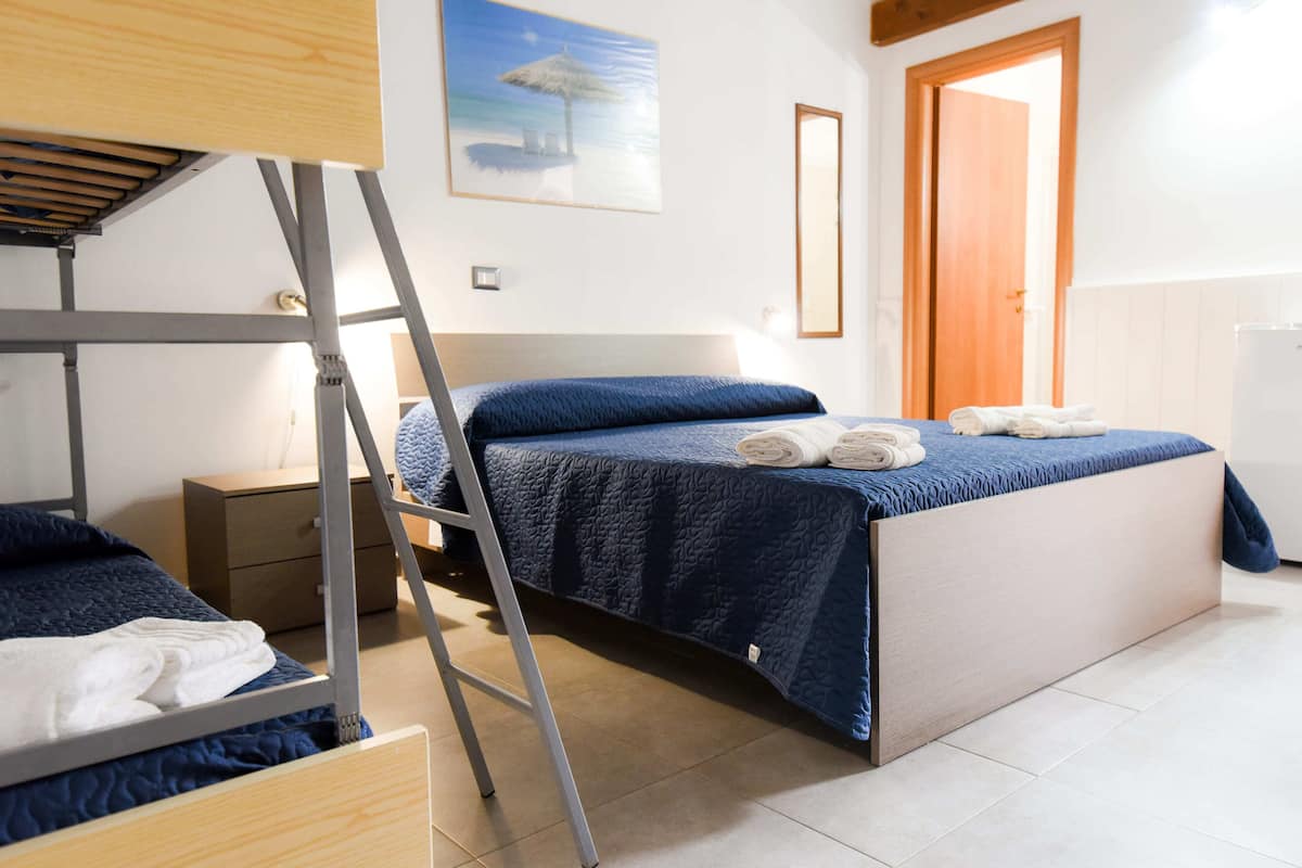 Een slaapkamer met stapelbedden, een ladder, een nachtkastje, een bed met een blauwe deken en een schilderij van een strand.