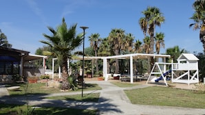 Un espacio al aire libre techado con un parque infantil, palmeras y un edificio con terraza cubierta.