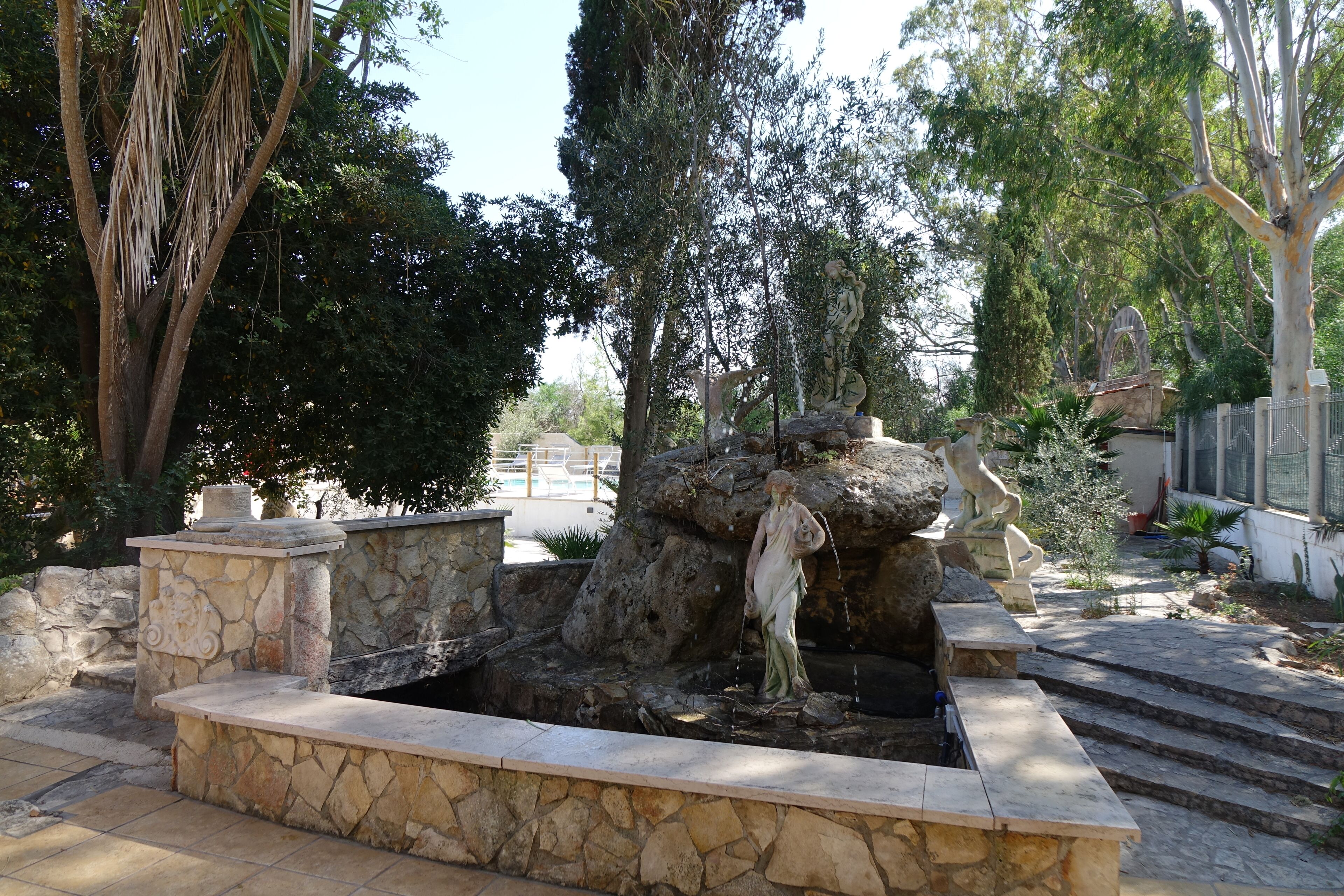 A stone fountain with a statue, surrounded by trees and steps leading up to a building.