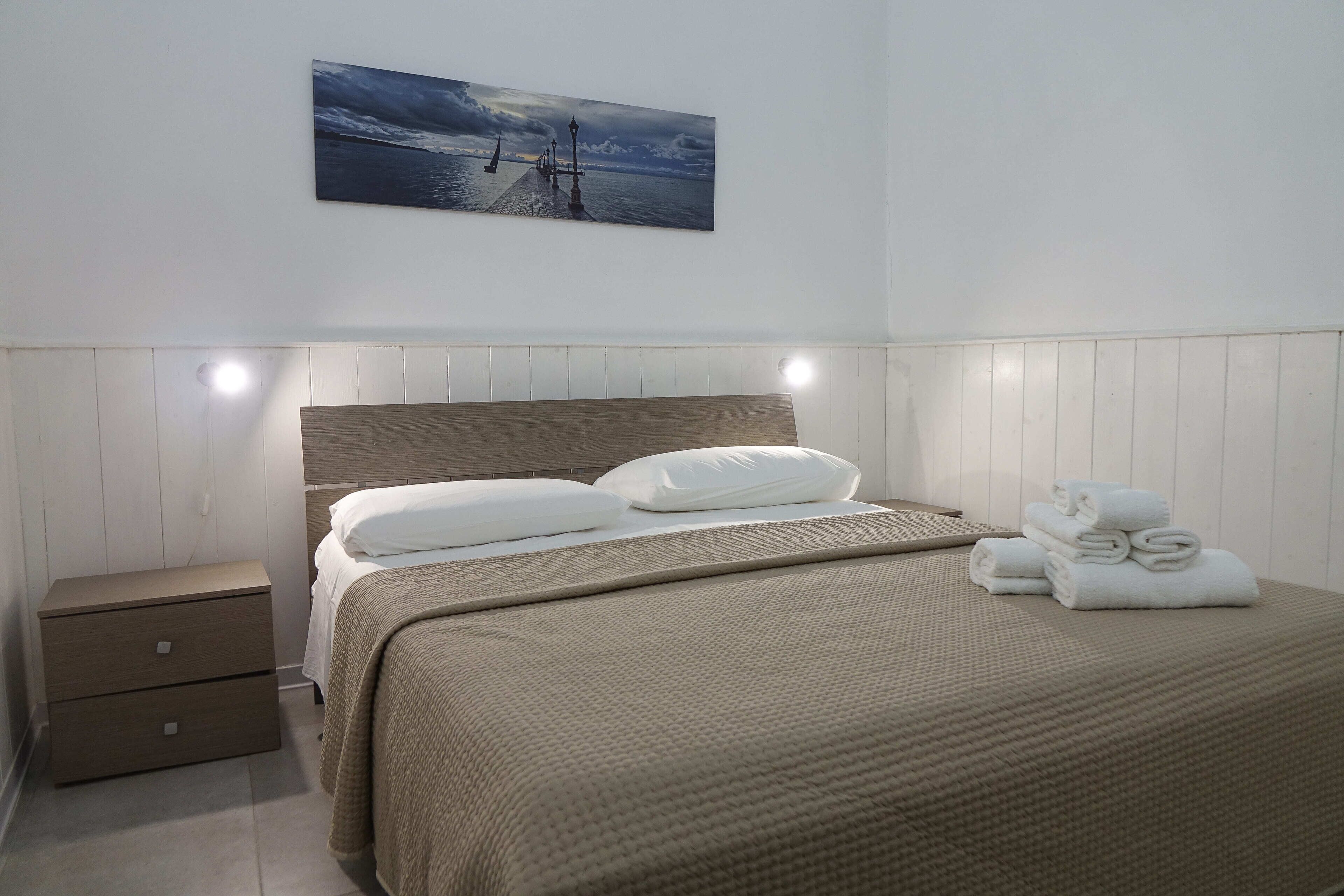 A neatly made bed with a headboard, a bedside table with drawers, and a wall-mounted painting of a pier.