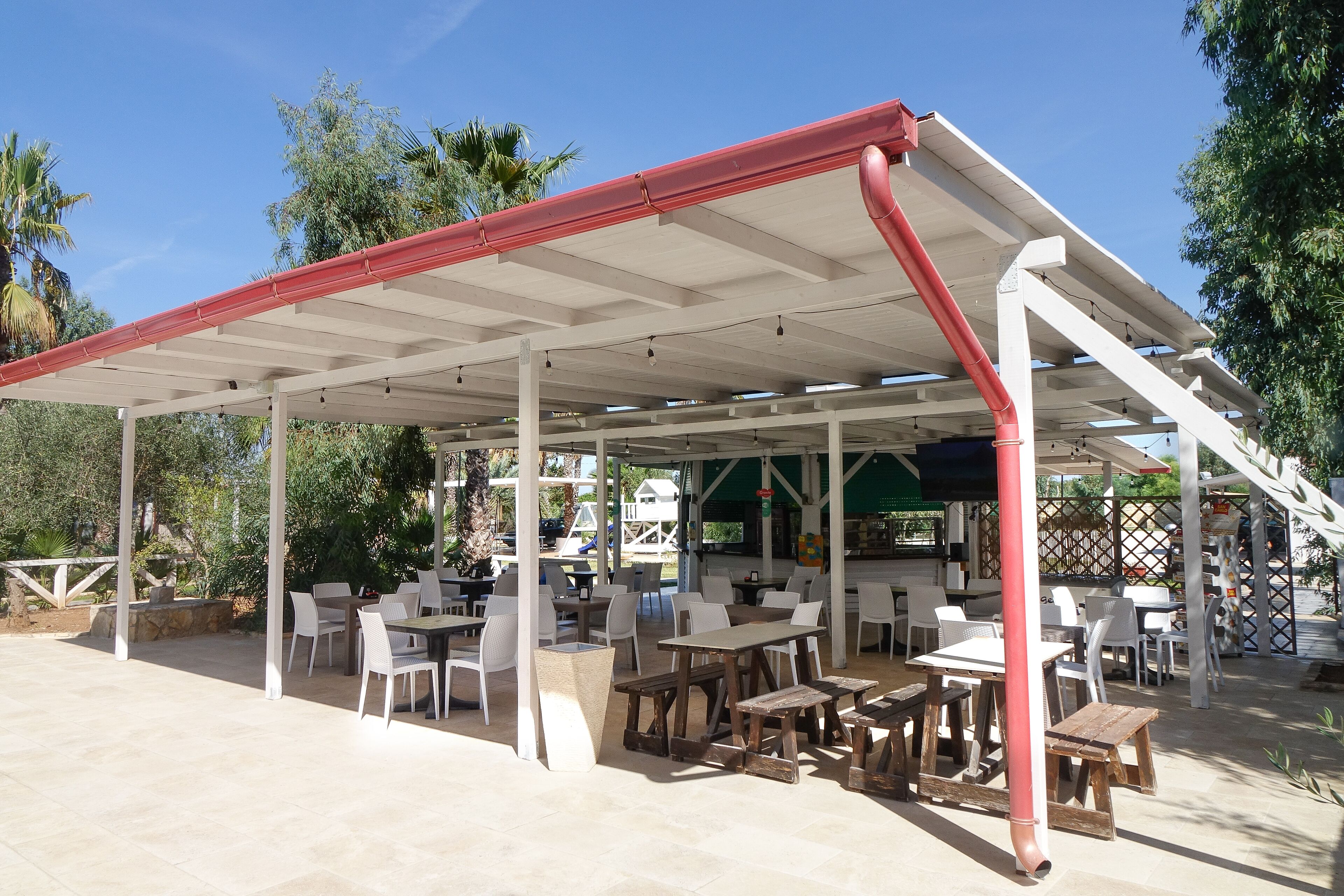 An outdoor patio area with white tables and chairs, a covered seating area with a red roof, and a view of the surrounding greenery.