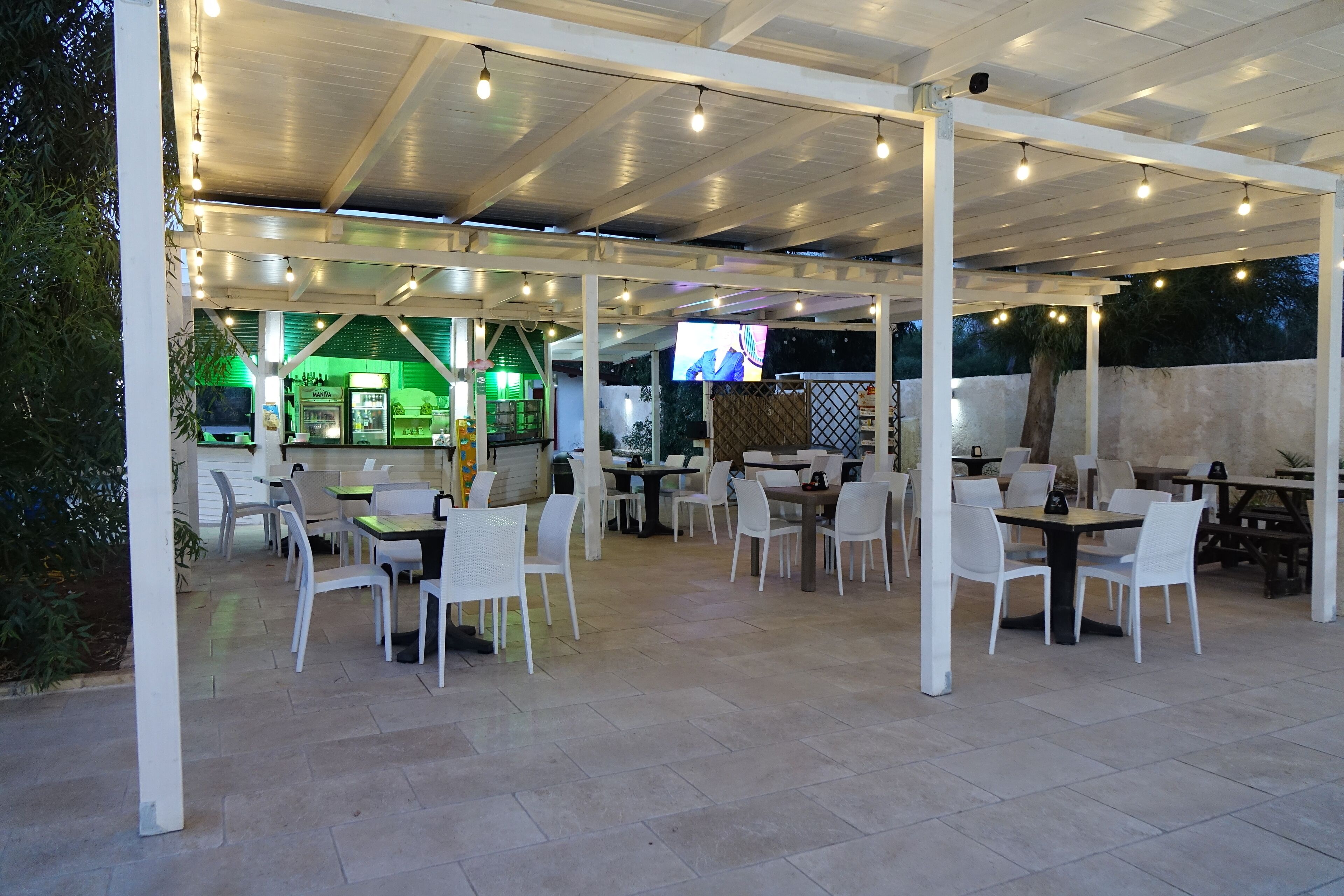 An outdoor patio area with tables and chairs, a green-lit bar area, and a television screen.