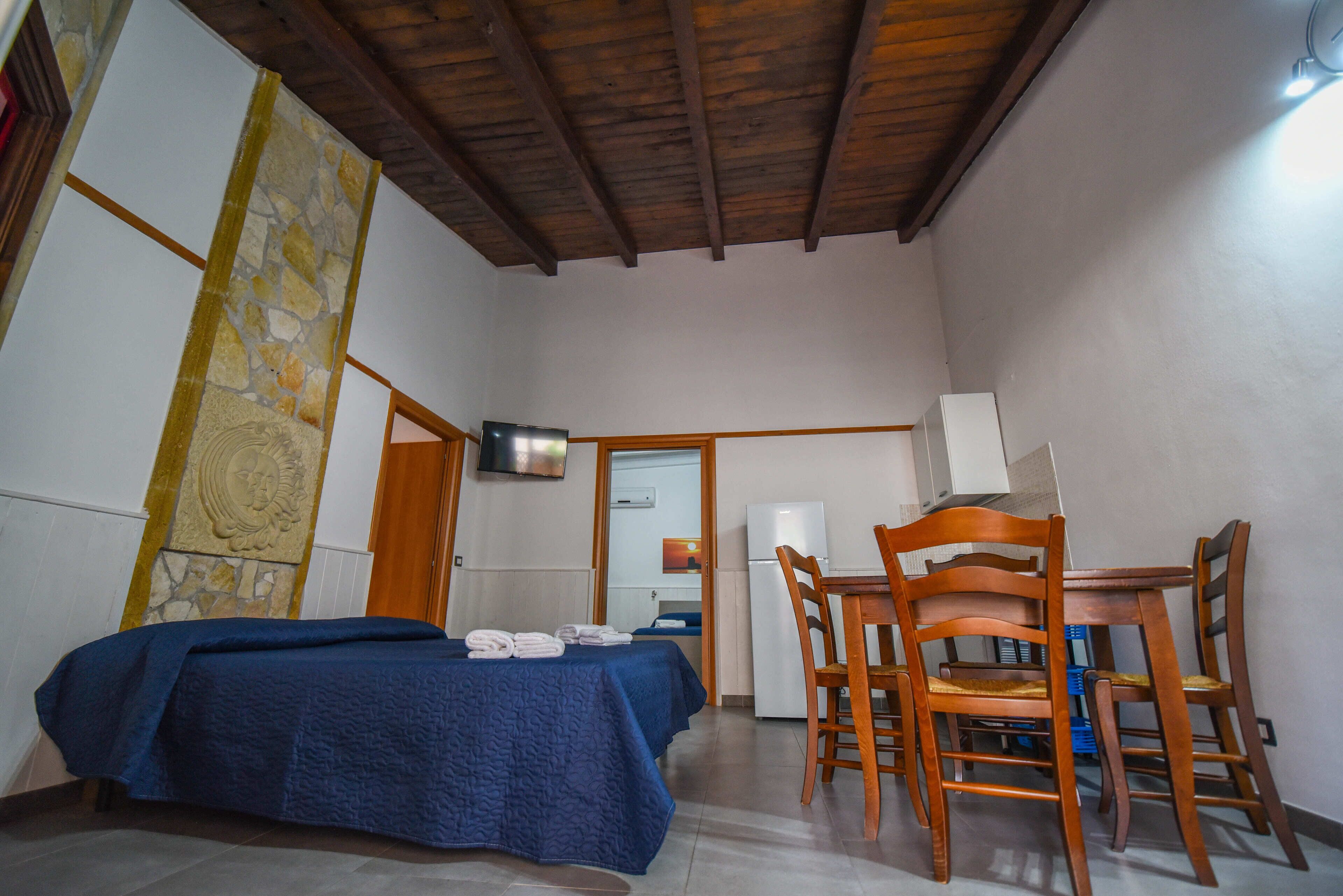 Una habitación con cama, una mesa de comedor de madera con sillas, un refrigerador y una pared de piedra con un adorno.