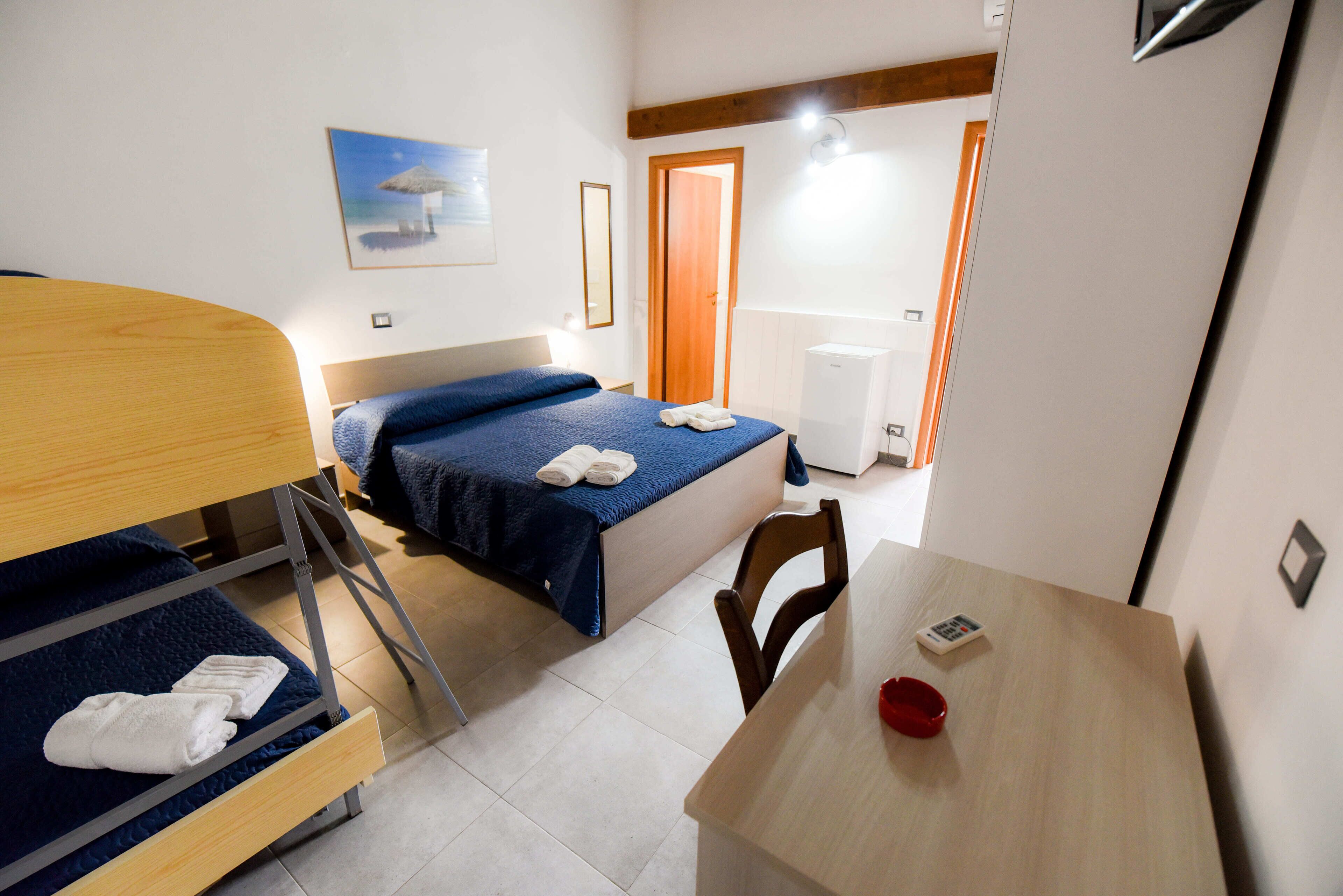 Ein Hotelzimmer mit Etagenbett, einem Einzelbett, einem Holztisch, einem Stuhl, einer kleinen roten Schale und einer Wandleuchte.