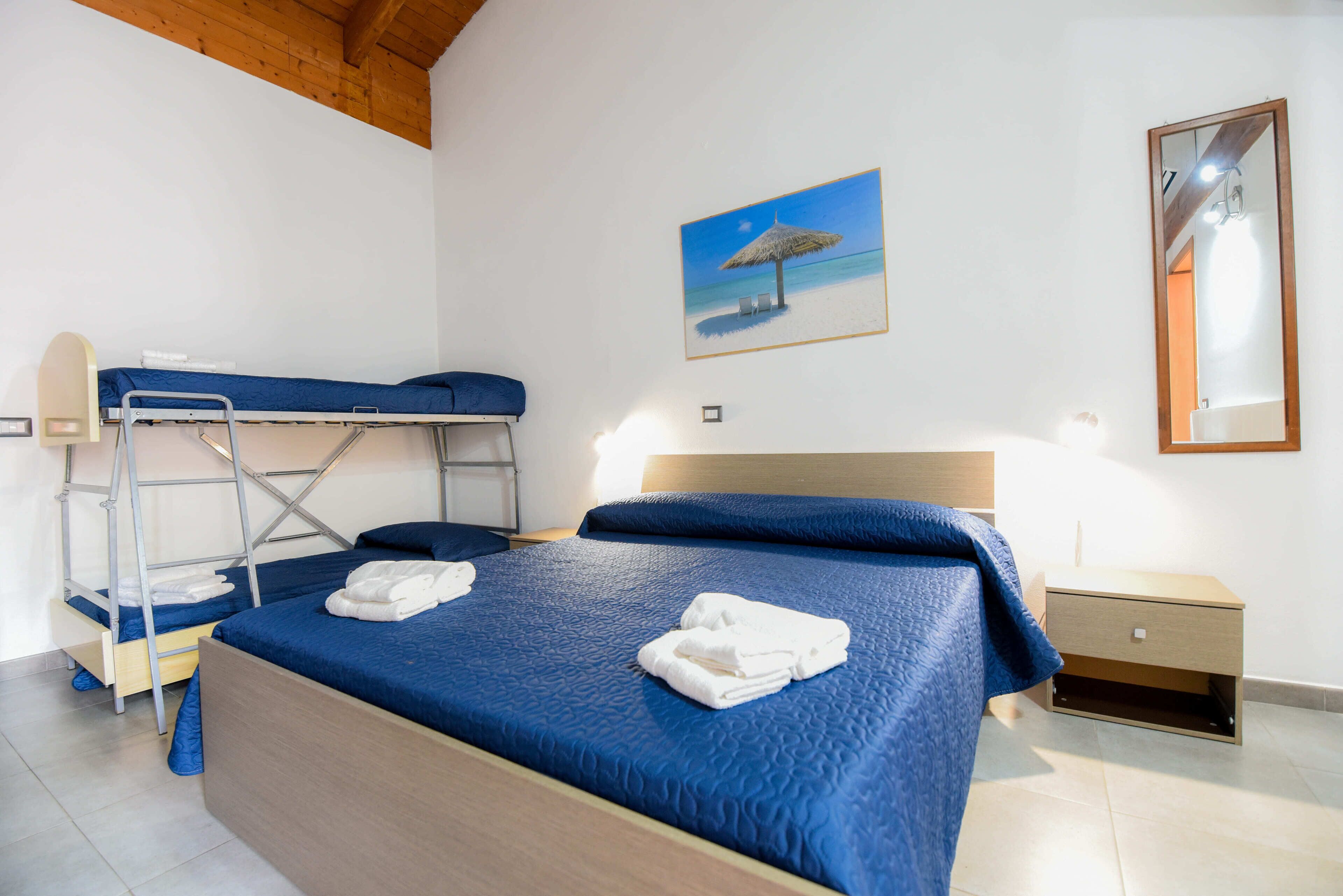 A room with bunk beds, a single bed with blue bedding, a small bedside table, and a painting of a beach scene.