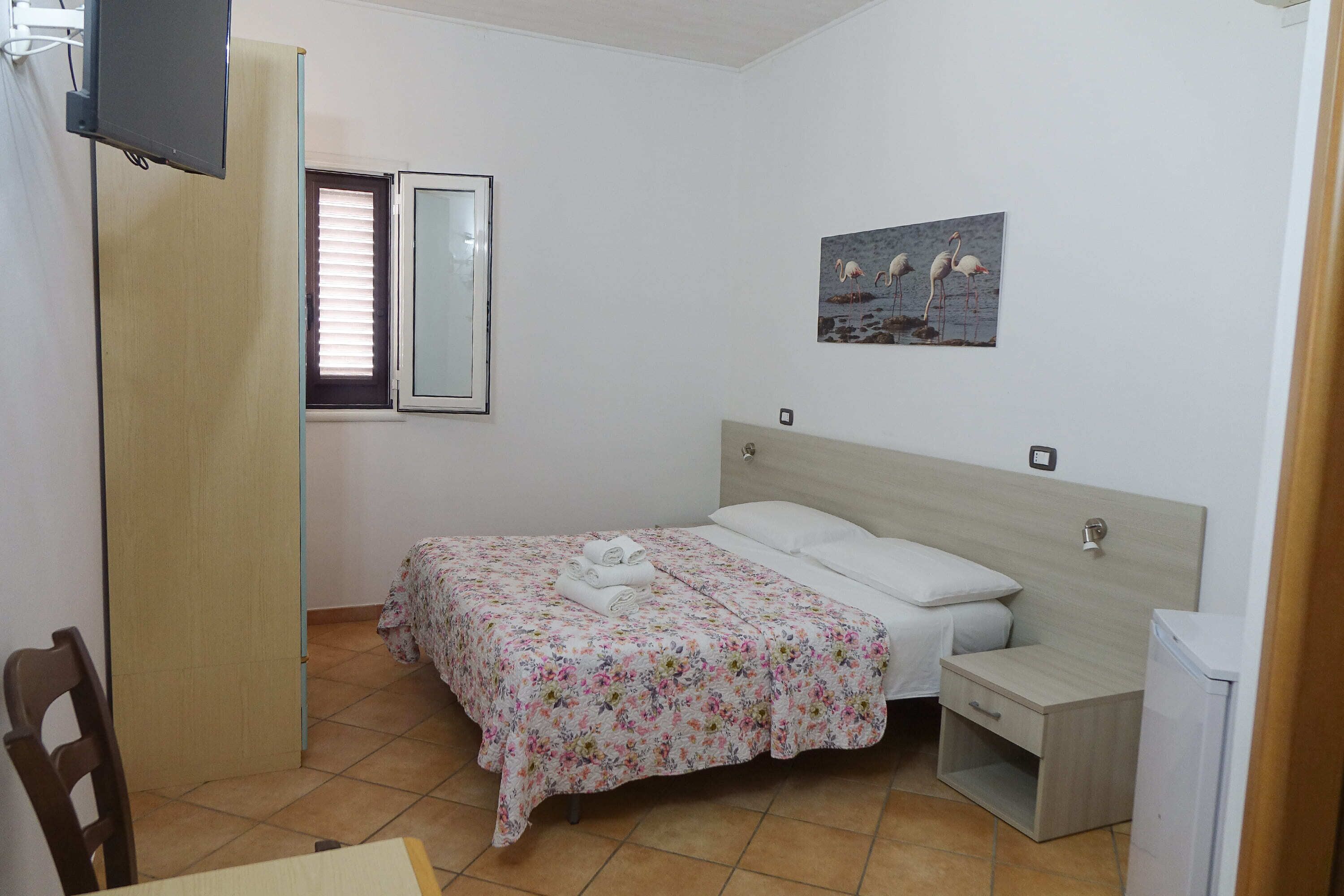 A bedroom with a bed, a bedside table, a chair, a TV, a window, and a painting on the wall.