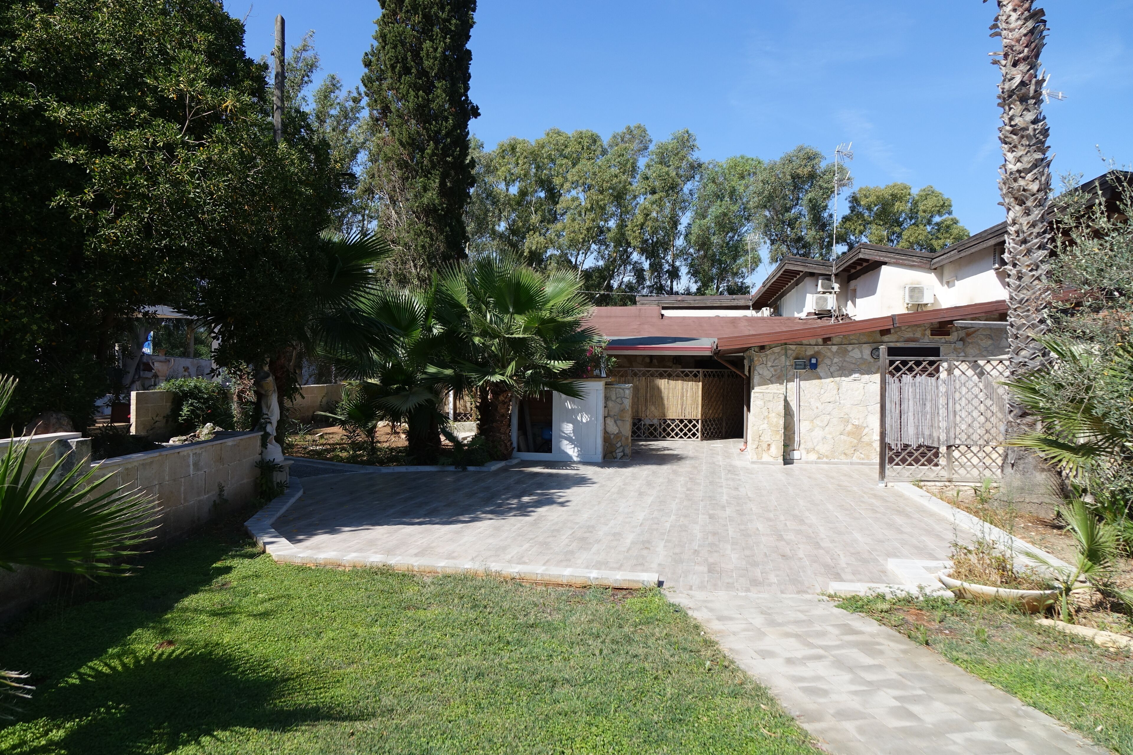 A residential property with a paved driveway, stone walls, and a fenced yard with palm trees.