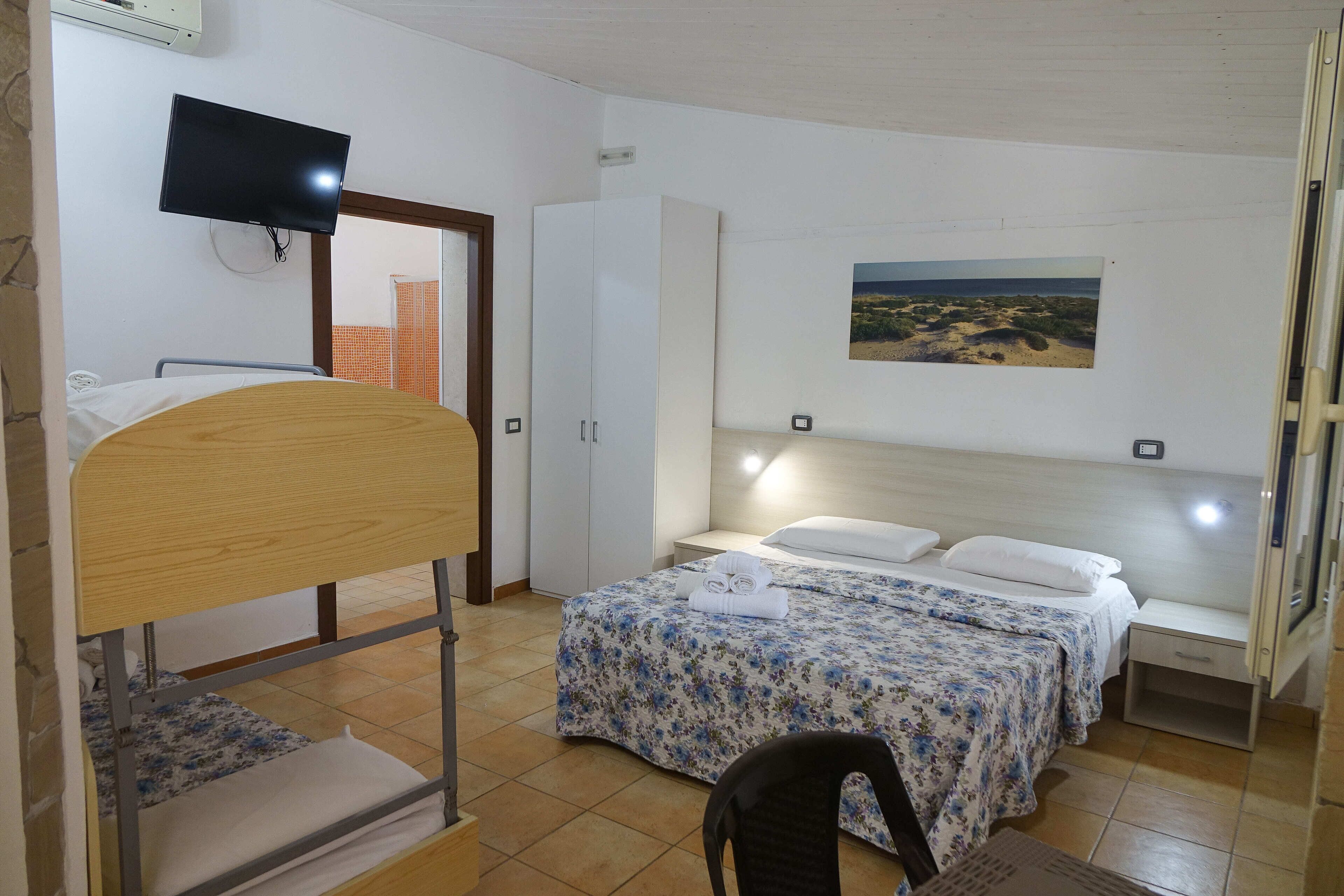A hotel room with a bunk bed, a single bed with floral bedding, a flat-screen TV mounted on the wall, and a painting of a beach scene.