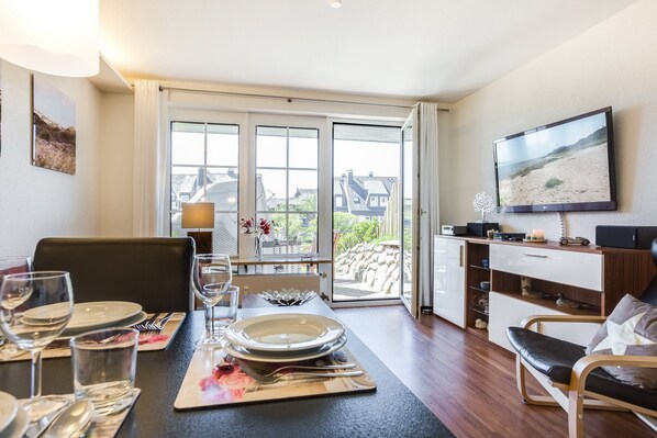 Un comedor con una mesa dispuesta para cuatro personas, un televisor en la pared y vistas al exterior a través de grandes ventanales.