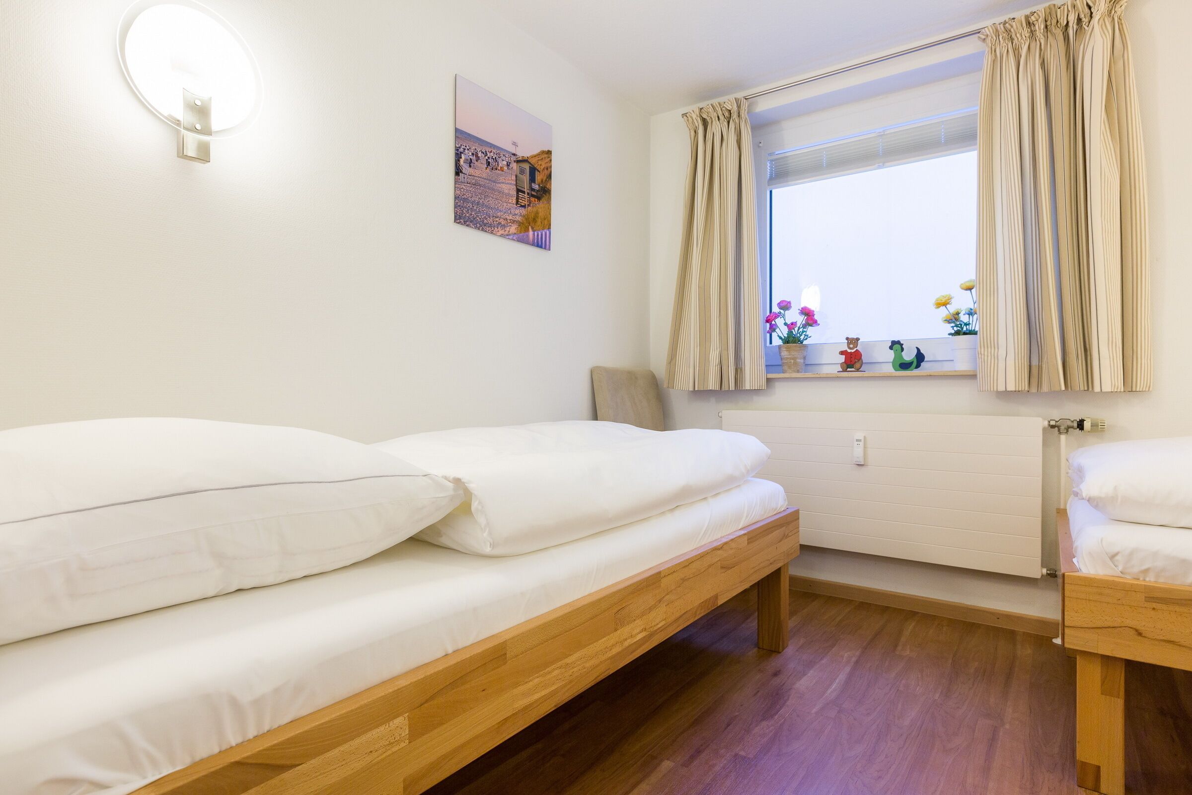 A small, clean room with a wooden bed, a window with curtains, a radiator, and a wall-mounted light fixture.