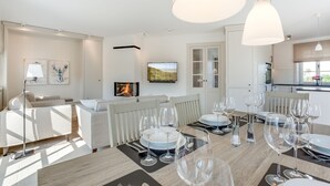 A modern living room with a dining area set for a meal, featuring a fireplace, white furniture, and a view of the outdoors.