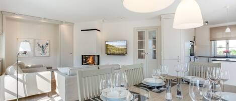 A modern living room with a dining area set for a meal, featuring a fireplace, white furniture, and a view of the outdoors.