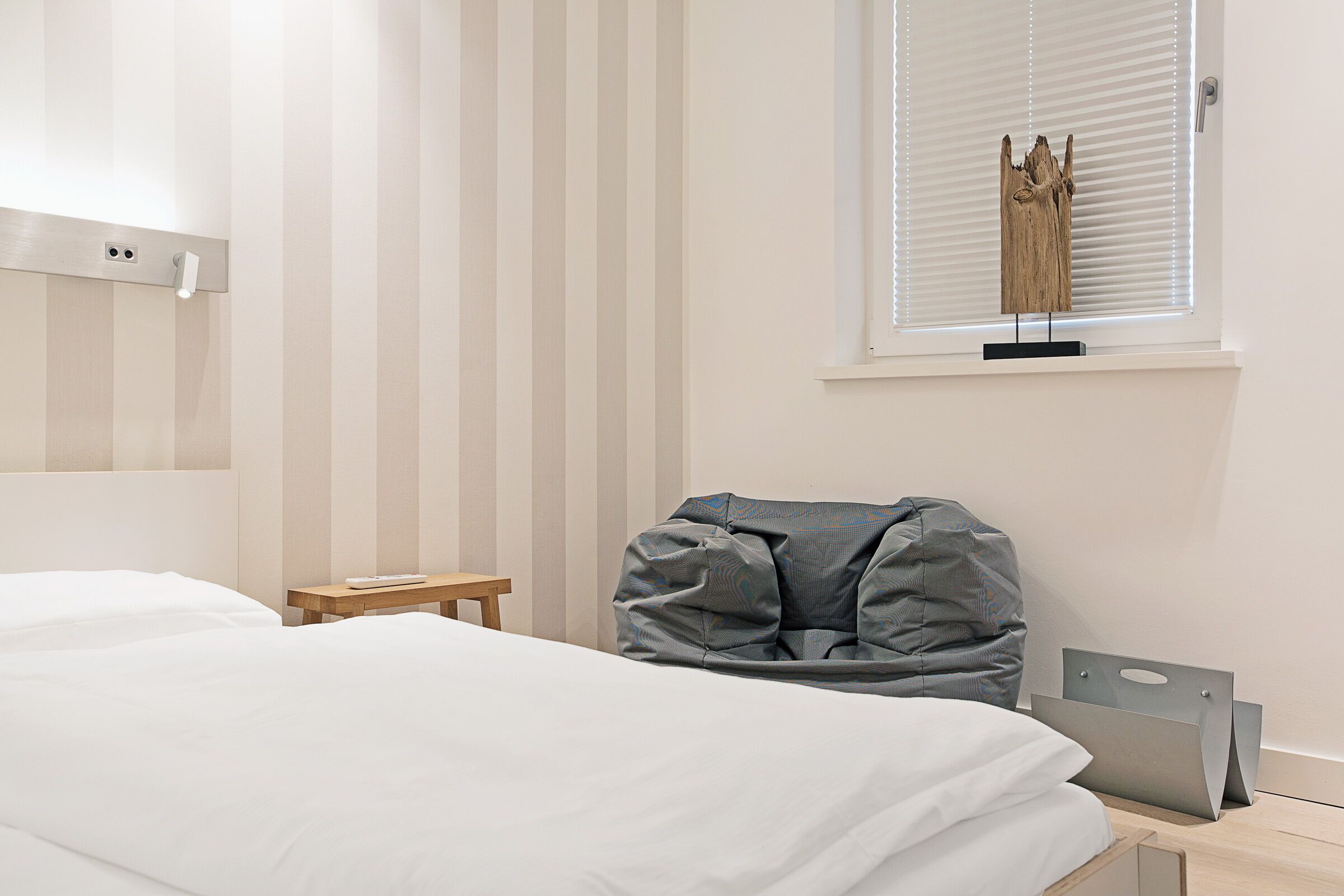 A bedroom with a bed, a small bedside table, a gray bean bag chair, and a window with blinds.