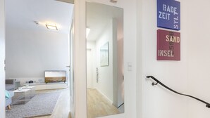 A modern interior with a hallway leading to a living area. A large mirror reflects the room, and there are two framed signs on the wall.