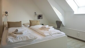 A neatly made bed with white linens and towels, a bedside lamp, and a wall-mounted artwork in a room with a sloped ceiling and a radiator.