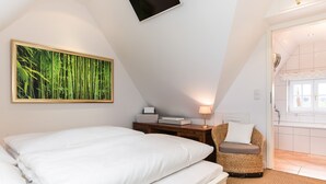 Een slaapkamer met een bed, nachtkastje, lamp, een rieten stoel, en een badkamer die door een geopende deur zichtbaar is.