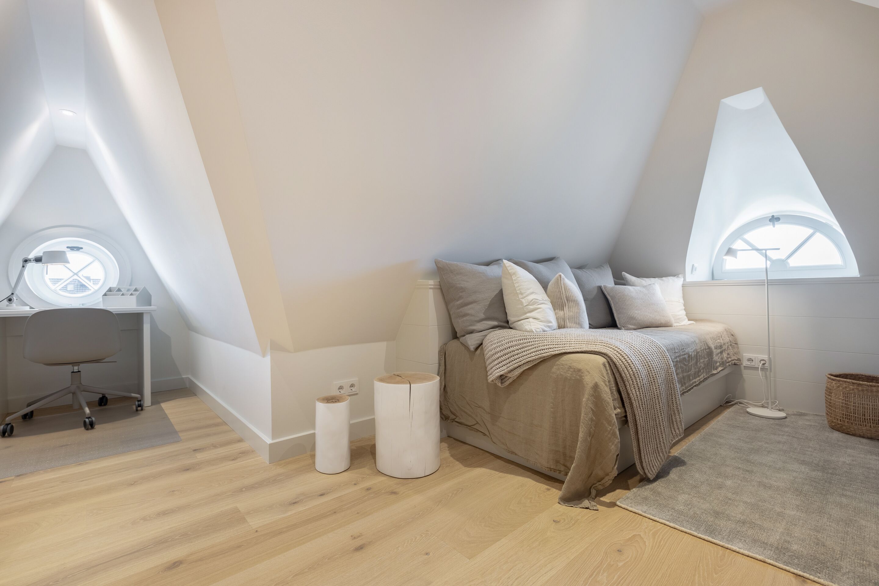 Ein modernes Schlafzimmer mit schrägverdeckter Decke, einem Rundfenster, einem Bett mit Kissen, einem Schreibtisch mit Stuhl und einem Nachttisch mit Lampe.