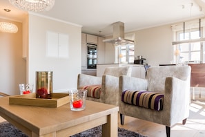 Una sala de estar moderna con una mesa de centro de madera, dos sillones estampados y una cocina al fondo.