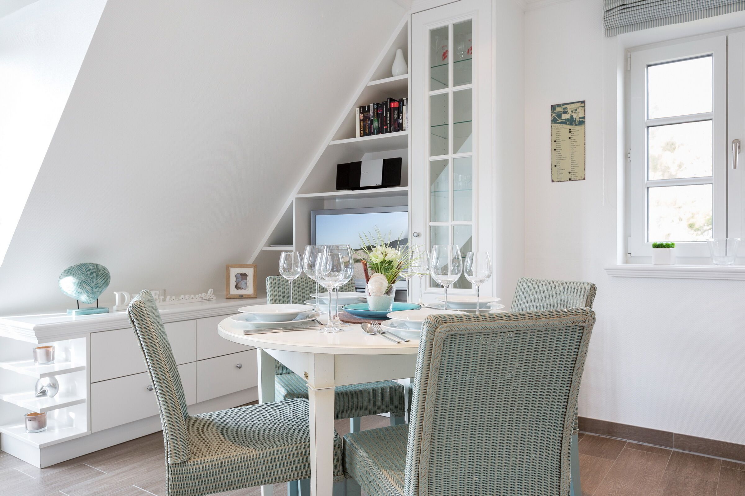 A dining area with a white table set for four, surrounded by wicker chairs. A white cabinet with glass doors is to the left, and a window with a view of greenery is to the right.