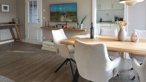 A modern dining area with a wooden table, white chairs, a television above the table, and a kitchen in the background. - Strandnaher State-of-the-art Luxus (Wenningstedt-Braderup (Sylt))