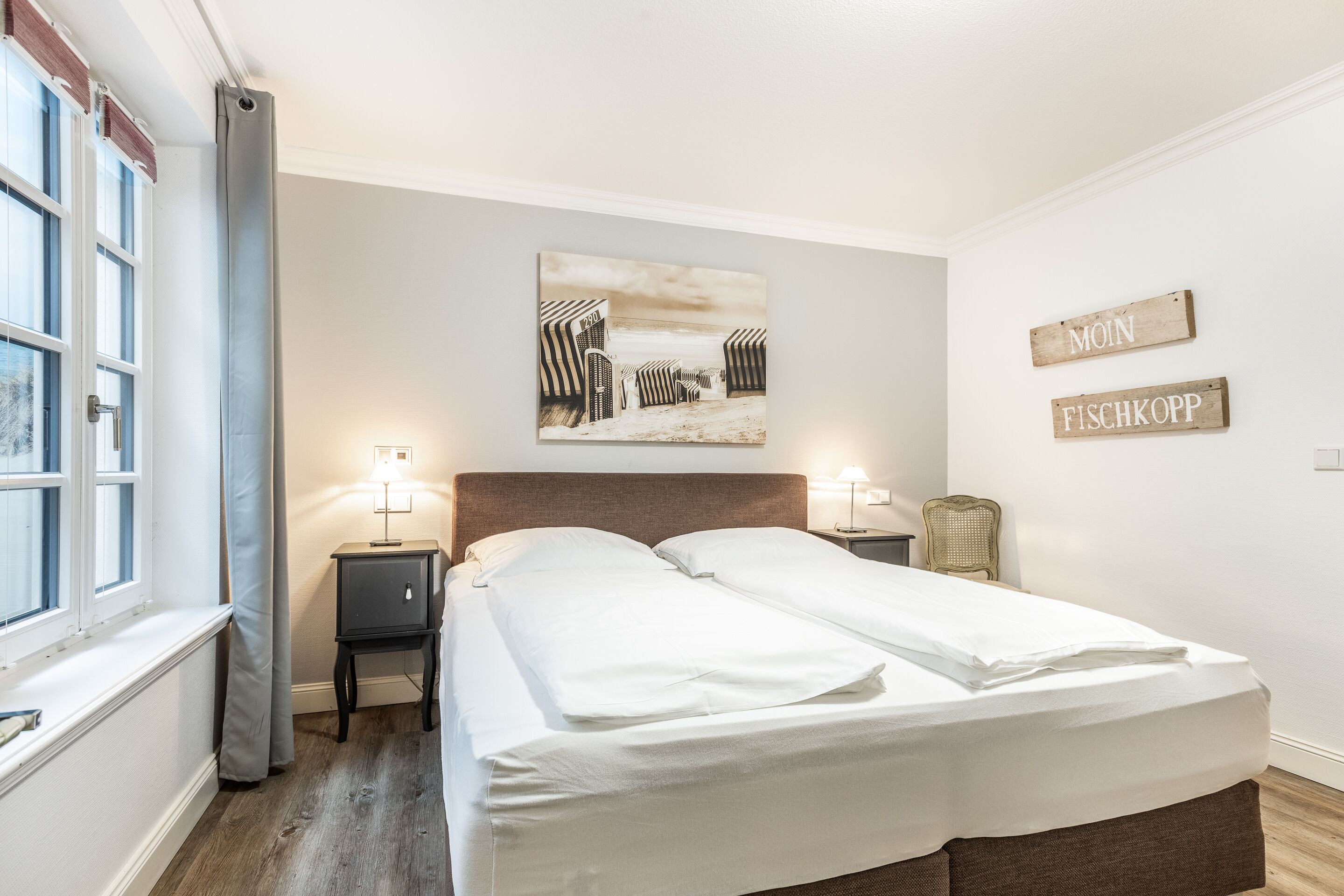 Un lit double avec du linge de lit blanc, une tête de lit, des tables de chevet avec des lampes, une fenêtre avec des rideaux et un tableau sur le thème de la plage accroché au mur.