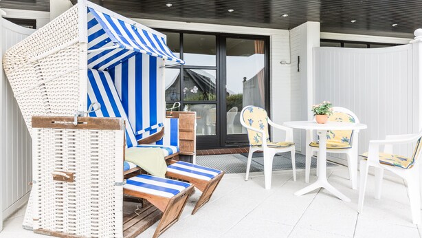 A covered outdoor seating area with wicker furniture and striped cushions, a white table with chairs, and a view of a modern building in the background. - Komfortables Appartement im Erd- und Untergeschoss, ca. 50 qm Nutzfläche (Wenningstedt-Braderup (Sylt))