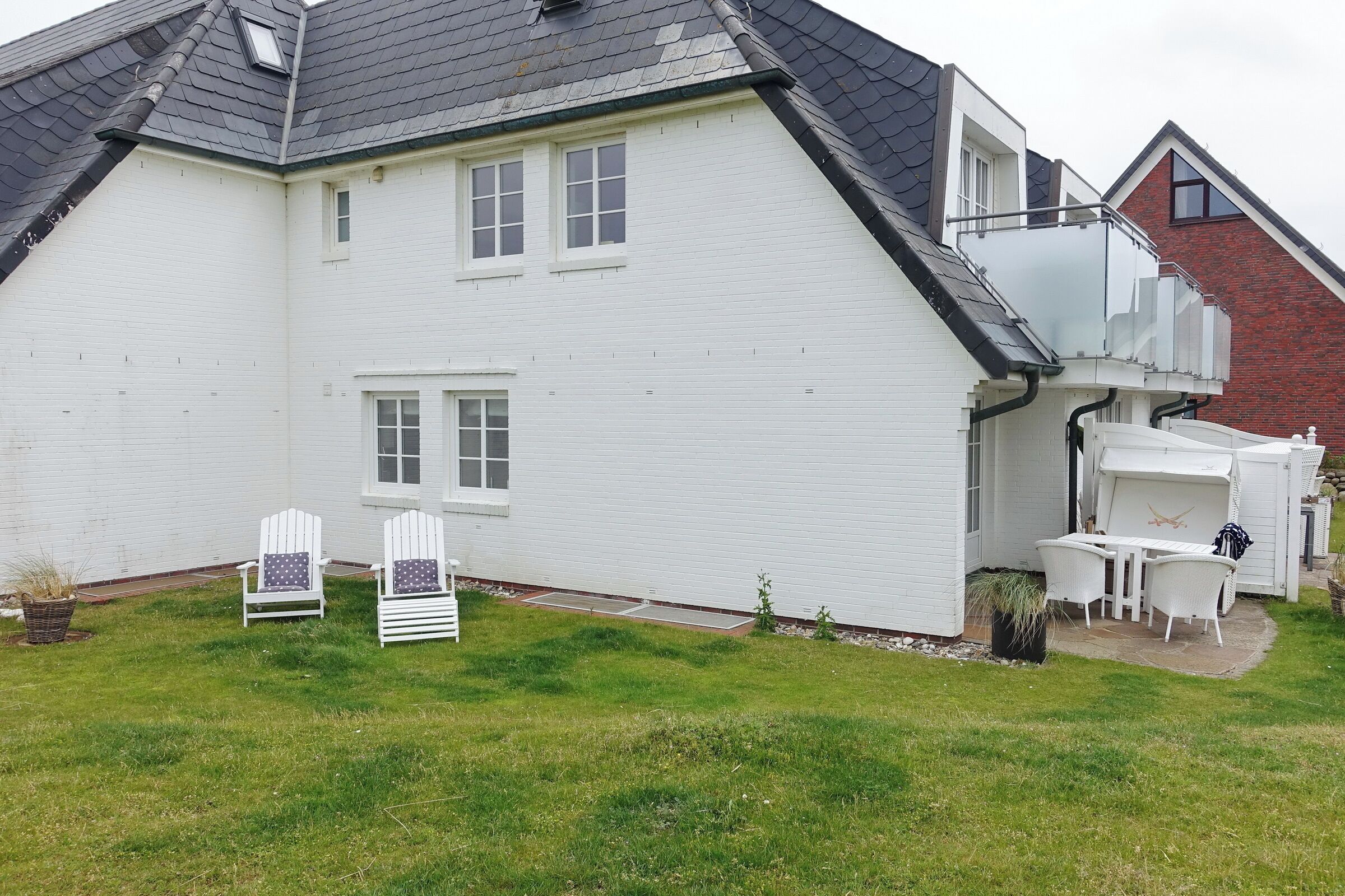 Una casa blanca con techo inclinado, un balcón y un pequeño patio con mobiliario exterior.