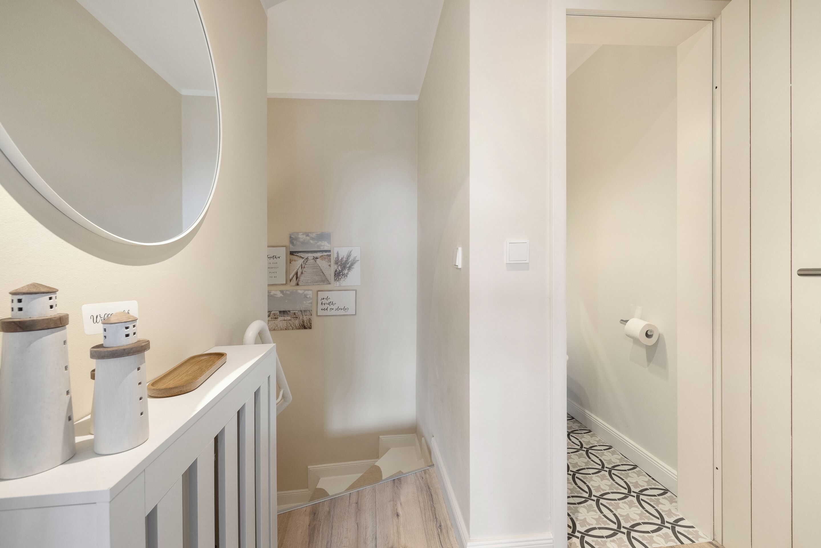 A modern bathroom with a white vanity, a round mirror, and decorative jars. A wooden tray is on the vanity, and there are framed pictures on the wall.