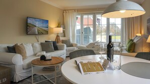 Ein helles Wohnzimmer mit einem weißen Sofa, einem runden Tisch mit einer Zeitschrift, einer Flasche und einem Glas.