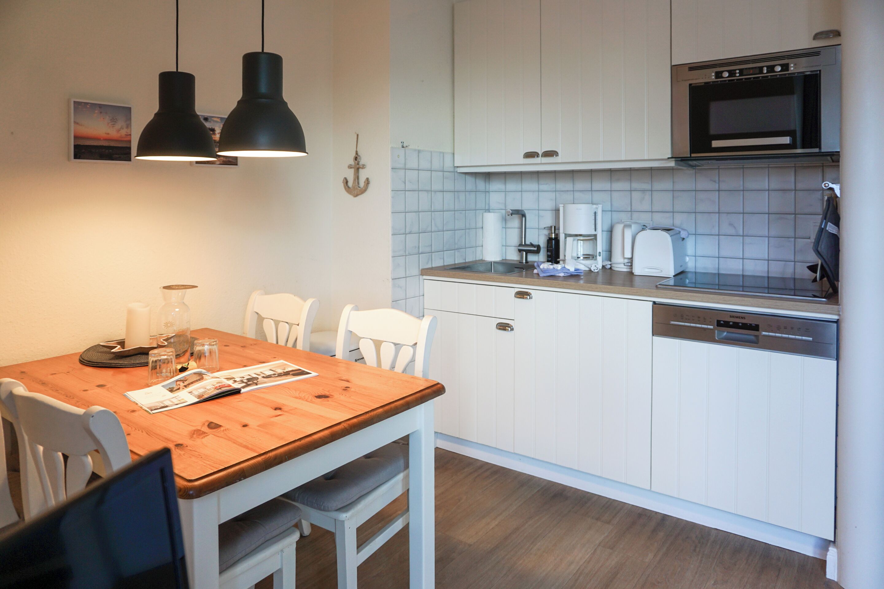 Cocina compacta con armarios blancos, microondas integrado y lavavajillas. Hay una mesa de comedor de madera con sillas y una pequeña isla con pileta.