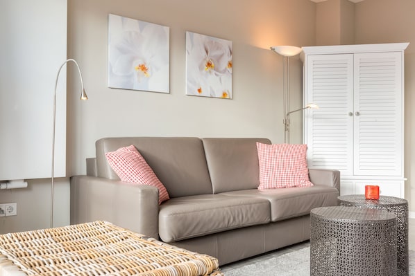 A modern living room with a beige sofa, two red and white checkered pillows, a woven ottoman, a round metal table, and two wall-mounted flower paintings.