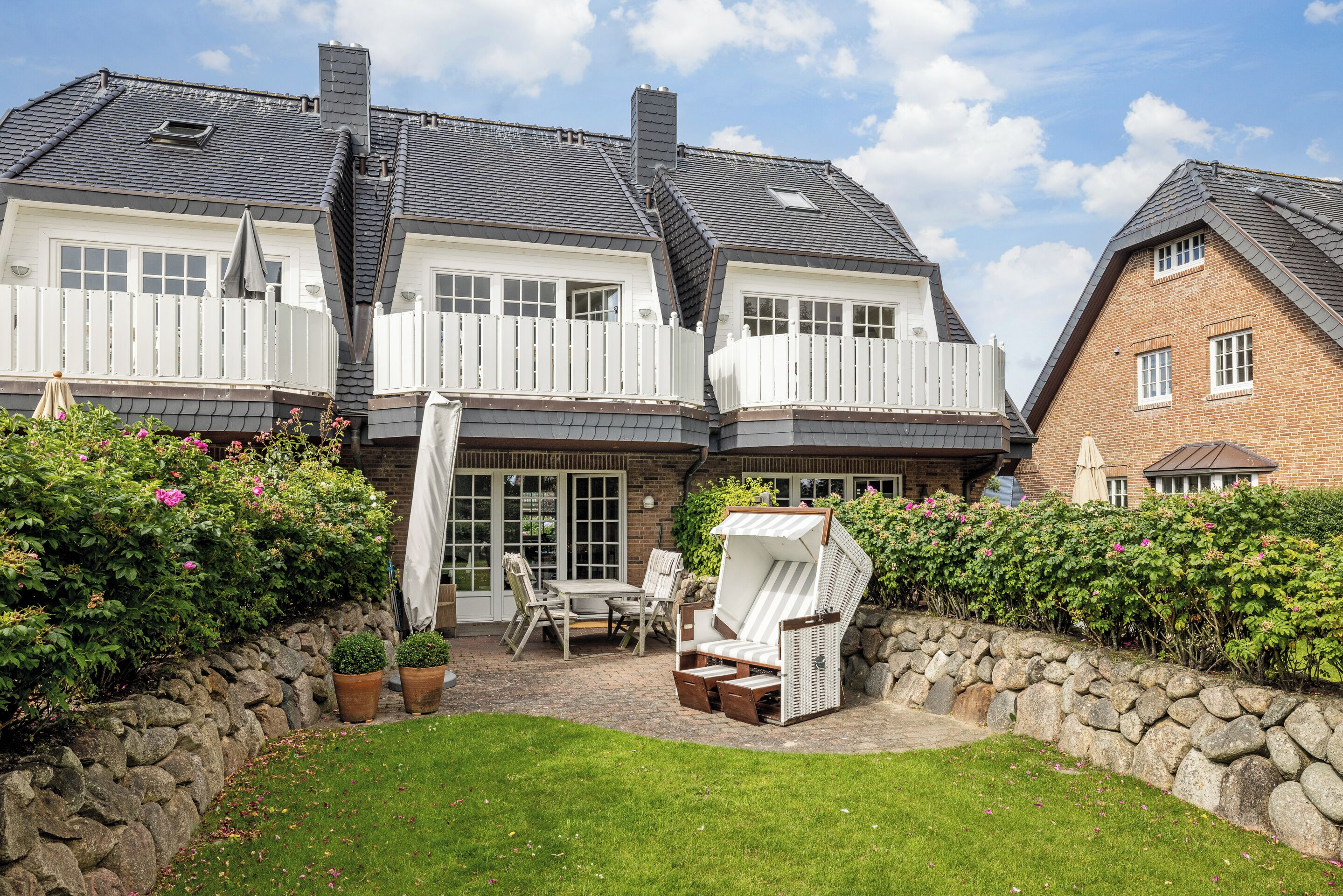 Een huis met twee verdiepingen, een stenen muur en een terras met tuinmeubilair.