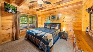Camera da letto in una baita di legno, con un letto, una ventola a soffitto, un comodino e una finestra con tende.