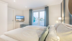 A hotel room with a large bed, a television, a view of the beach, and a window with curtains. - Düne 22 - Wohnen Unter Reet (Wenningstedt-Braderup (Sylt))