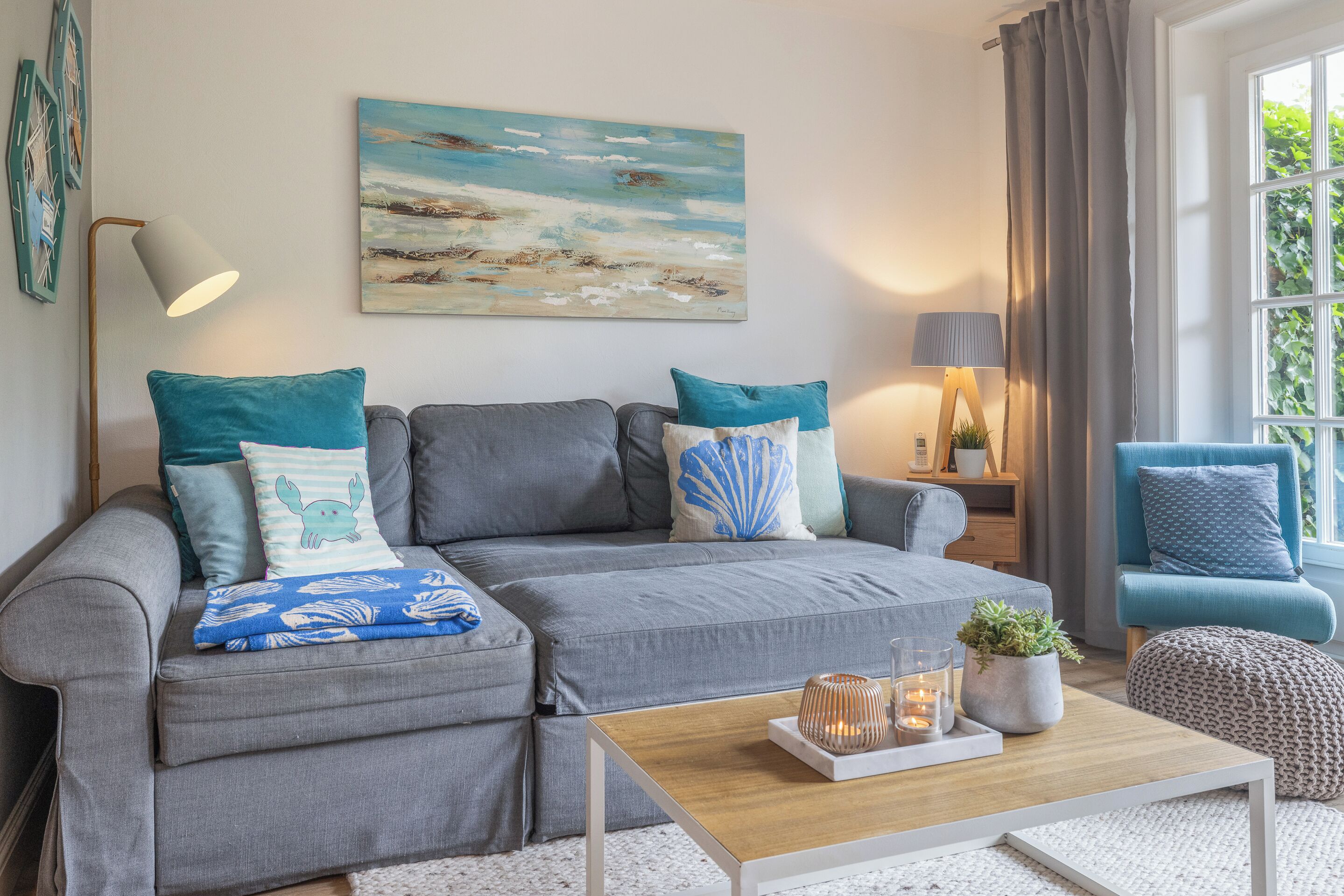 Un salon moderne avec un canapé gris, des coussins turquoise, une table basse en bois et un tableau représentant une scène de plage.