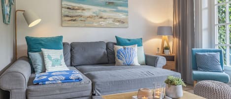 Un salon moderne avec un canapé gris, des coussins turquoise, une table basse en bois et un tableau représentant une scène de plage.